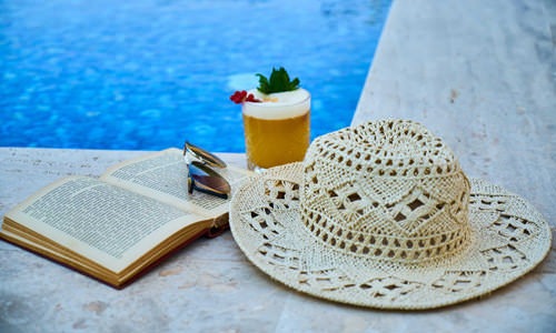 sun hat, open book and tropical drink near pool